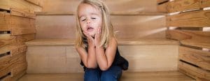 Adorable blond girl sitting on wooden stairs with hands on her cheeks and eyes closed