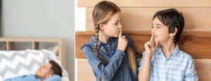 Siblings hold their fingers over their mouth to say shhhh while dad sleeps on the couch