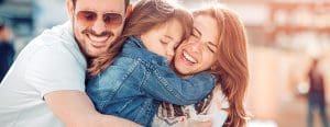 Parents squeeze hug their daughter in between them with big smiles